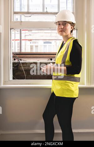 Vertical photo of a chartered civil engineer woman using a calculator in front of a window in an indoor construction site, wears hard hat, yellow ppe Stock Photo