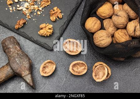 Walnuts in a reusable bag. Hammer, nut shells on the table and nut kernels on stone board. Black background. Stock Photo