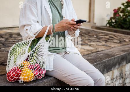 Woman with reusable bag sitting on wall and using smart phone in city. Resting after shopping Stock Photo