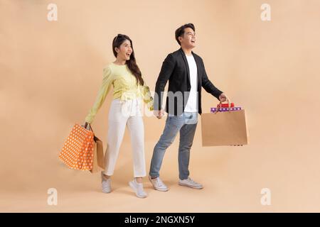 Happy smiling Asian couple shoppers holding a bunch of shopping bags while walking forward - Boyfriend and girlfriend shopping - with empty copy space - in studio full length Stock Photo