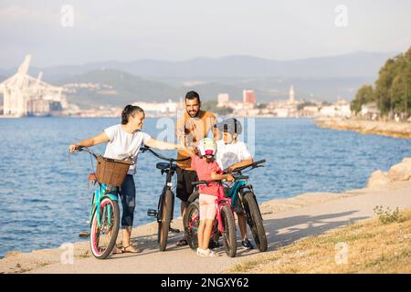 Caucasian family team, cheerful mother, father, and children, giving high five, standing with bikes on the seashore Stock Photo