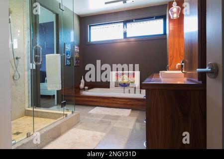 Walnut wood vanity and base with deep white acrylic bathtub, clear glass shower stall in en suite with grey and white marble floor inside home. Stock Photo