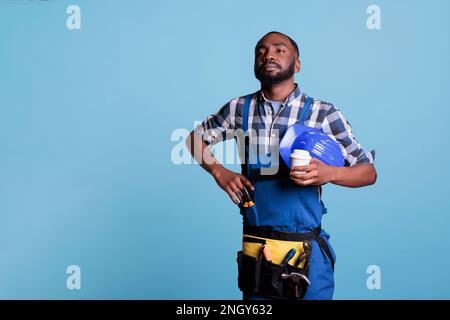 African american builder looking into the distance while resting with a cup of coffee in studio shot against blue background. Construction employee resting before continuing work day. Stock Photo