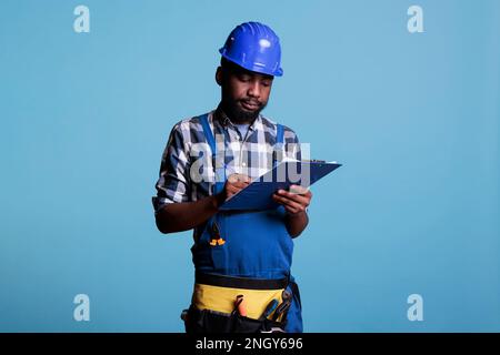 Construction supervisor writing on clipboard supervising building renovation work. Construction worker taking notes on sheet of paper to check job is perfect, studio shot against blue background. Stock Photo