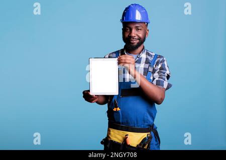 Portrait of contractor with digital tablet with empty white screen, showing it to camera standing in studio with blue background. Construction employee wearing coveralls and hard hat holding device. Stock Photo