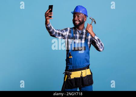 African american contractor taking selfie with mobile phone holding hammer against blue background in studio shot. Construction worker in work uniform and tool belt looking happy. Stock Photo