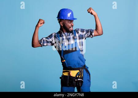 Funny african american construction worker in dungarees showing arm muscles in front of the camera in studio shot against blue background. Dressed in work uniform wearing tool belt. Stock Photo