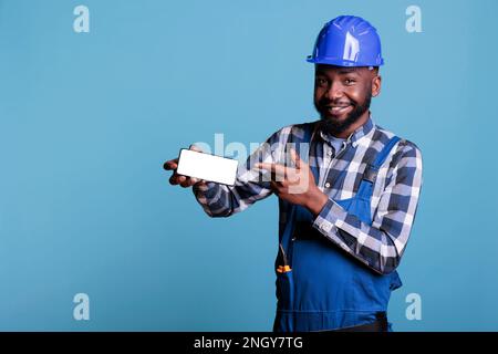 African american construction worker in protective helmet pointing finger at cell phone with empty screen isolated on blue background. Builder in studio shot holding advertising blank display. Stock Photo