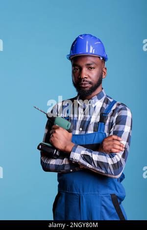 African american builder holds electric cordless drill while looking seriously into camera. Contractor holding work tool with confidence, expert in construction and renovations. Stock Photo