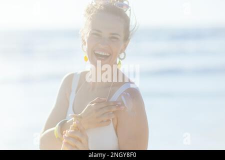 Portrait of happy elegant middle aged woman in white beachwear at the beach using sunscreen. Stock Photo