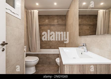 Small bathroom with a one-piece resin sink on oak furniture with drawers, a bathtub with a white plastic curtain and porcelain toilets Stock Photo