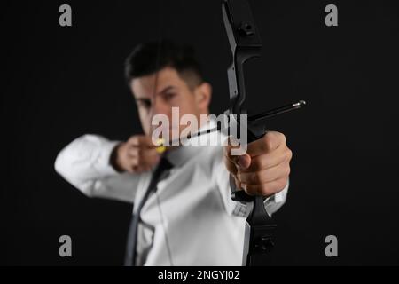 Businessman with bow and arrow practicing archery against black background, focus on hand Stock Photo