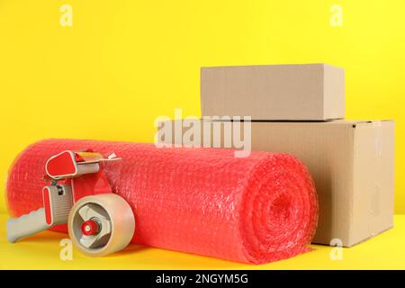 Bubble wrap roll, tape dispenser and cardboard boxes on yellow background Stock Photo