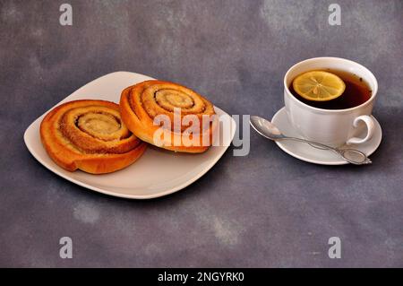 A plate with two spiral cinnamon rolls and a cup of hot tea with a slice of lemon on a gray background. Close-up. Stock Photo