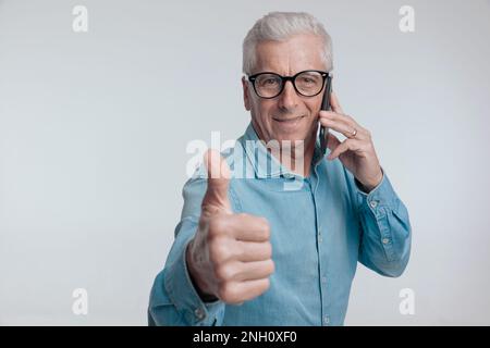 portrait of old man talking on the phone and making thumbs up gesture in front of grey background Stock Photo