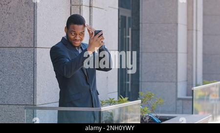 African American office worker man businessman standing on balcony holding smartphone looking in mobile screen reflection check hairstyle take picture Stock Photo