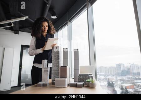 People, global business, company worker concept. Portrait of smiling lady with curly hair in formal clothes using tablet, posing in front of building complex prototype project of 3d city model. Stock Photo