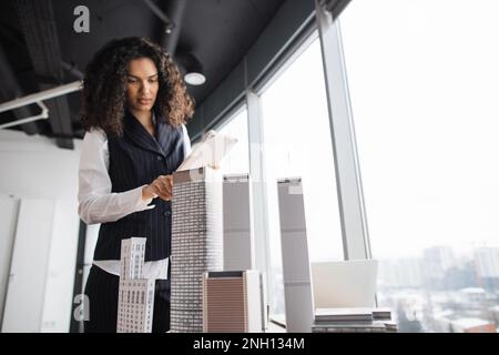 People, global business, company worker concept. Portrait of smiling lady with curly hair in formal clothes using tablet, posing in front of building complex prototype project of 3d city model. Stock Photo