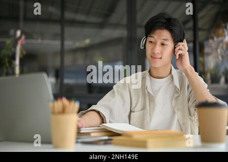 Happy and handsome young Asian man listening to music through wireless headphones while using laptop and remote working at the cafe. Stock Photo