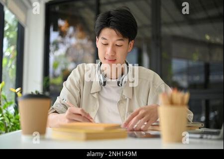 Smart and focused young Asian male college student is in the coffee shop doing homework, reading a book, preparing for an exam. Stock Photo
