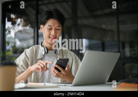 Handsome and focused young Asian male college student or freelancer in casual clothes using his smartphone at the table in the coffee shop. Stock Photo
