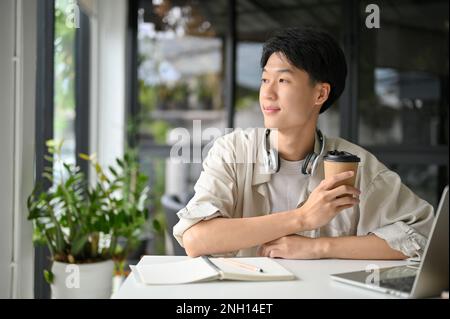 Relaxed and happy young Asian male college student or freelancer remote working at the coffee shop, sipping coffee, pensively thinking something while Stock Photo