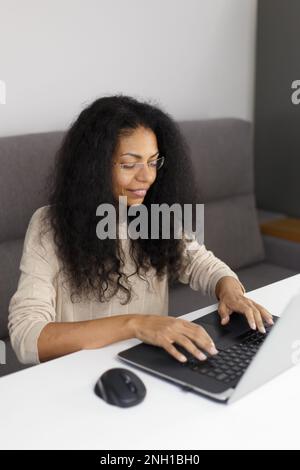 Cheerful black woman in glasses sitting behind desk and typing text on computer. Young adult POC female working on modern laptop at home Stock Photo