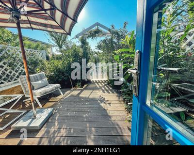 Open the glass door to a cozy patio area. There is a striped beach umbrella, a table and a bench with soft cushions and a beautiful green garden. Ahea Stock Photo