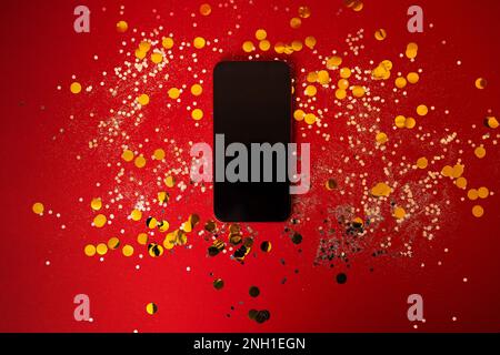 New mobile phone lies on a red background among silver sequins and round gold confetti. Phone with blank screen. Great mock up for a holiday theme. Cl Stock Photo