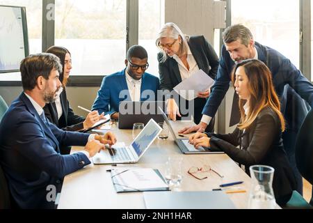 Group of multiethnic coworkers in formal clothes gathering at table while using laptops and discussing project details working on business strategy in Stock Photo