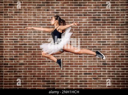 Side view full body of young ballerina in white tutu and black sneakers jumping against brick wall Stock Photo