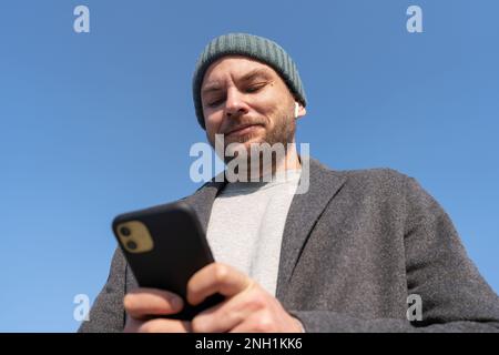 Man in hat and coat using his mobile phone and smiling while standing outdoors against blue sky. Stock Photo