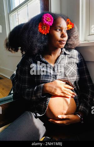 A pregnant woman sitting, leaning on mirror looks out window Stock Photo