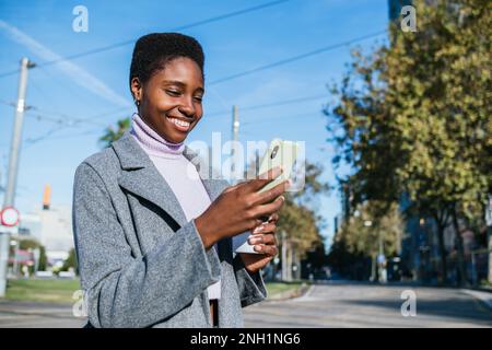 Happy black woman in stylish coat and sweater with cup of coffee smiling and scrolling news on smartphone while standing against blue sky and street of Barcelona, Spain Stock Photo