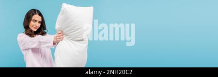 brunette young woman in pajamas holding white pillow isolated on blue, banner,stock image Stock Photo