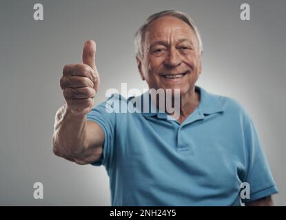 Id do this day all over again happily. a happy elderly man giving the thumbs up in a studio against a grey background. Stock Photo