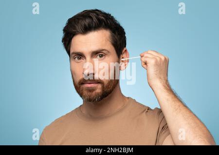 Bearded man cleaning ears, using cotton bud for wax removal, grooming hygiene and self care, blue background Stock Photo