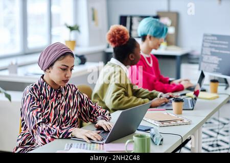 Diverse group of creative young people sitting in row while working in office focus on Muslim woman using laptop in foreground Stock Photo
