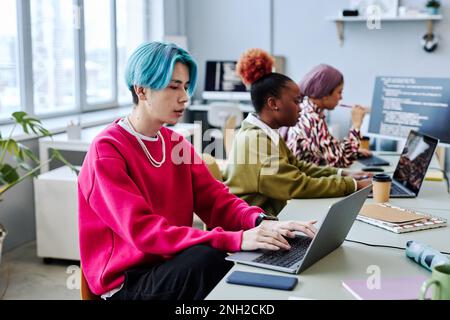 Diverse team of gen Z software developers working in office focus on Asian young man with colored hair using computer in foreground Stock Photo