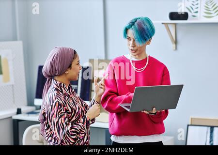 Waist up portrait of creative gen Z team discussing project while standing in office and holding laptop Stock Photo