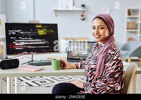 Portrait of Middle Eastern young woman working as software programmer and smiling at camera at workplace with code screens, copy space Stock Photo
