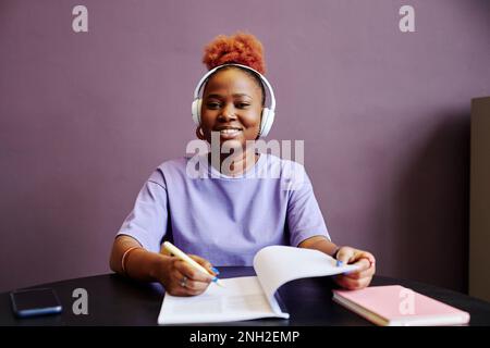 Minimal portrait of smiling black woman wearing headphones and taking notes in minimal purple interior Stock Photo