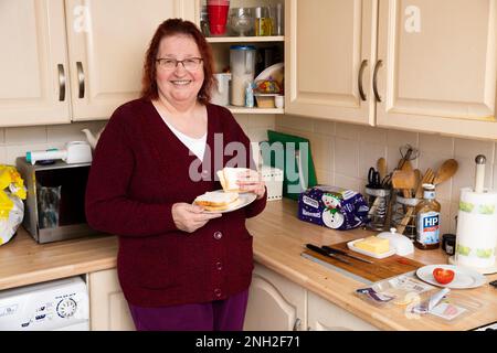 Older female standing in her kitchen having a sandwich. Manchester. United Kingdom. Stock Photo