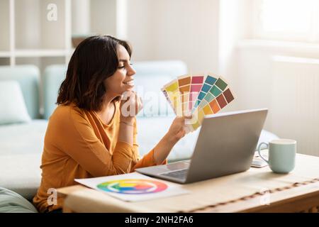 Middle Eastern Female Freelancer Holding Color Swatches And Using Laptop At Home Stock Photo