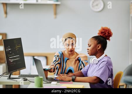 Portrait of two ethnic young women using computer while working on software development project together, senior specialist reviewing code Stock Photo