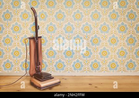 Retro styled image of a vintage brown vacuum cleaner in a room with floral wallpaper and wooden floor Stock Photo