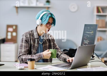 Portrait of young man with blue hair using computer in office and wearing headphones while writing code Stock Photo