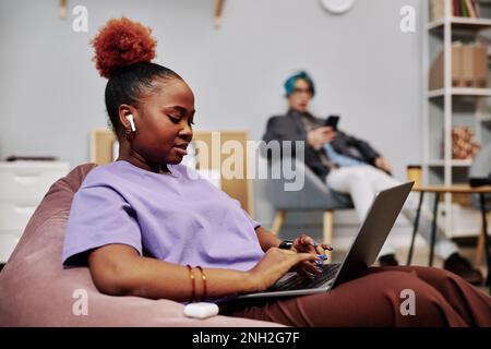 Side view portrait of young black woman using laptop in office lounge with wireless earphones Stock Photo