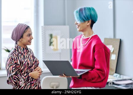 Side view portrait of two gen Z young people chatting while standing in office and holding laptop Stock Photo