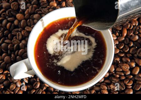 Pouring black coffee into white ceramic cup. Top view. Close up. Hot coffee drink pour into cup from shiny moka pot coffee maker. Roasted coffee beans Stock Photo
